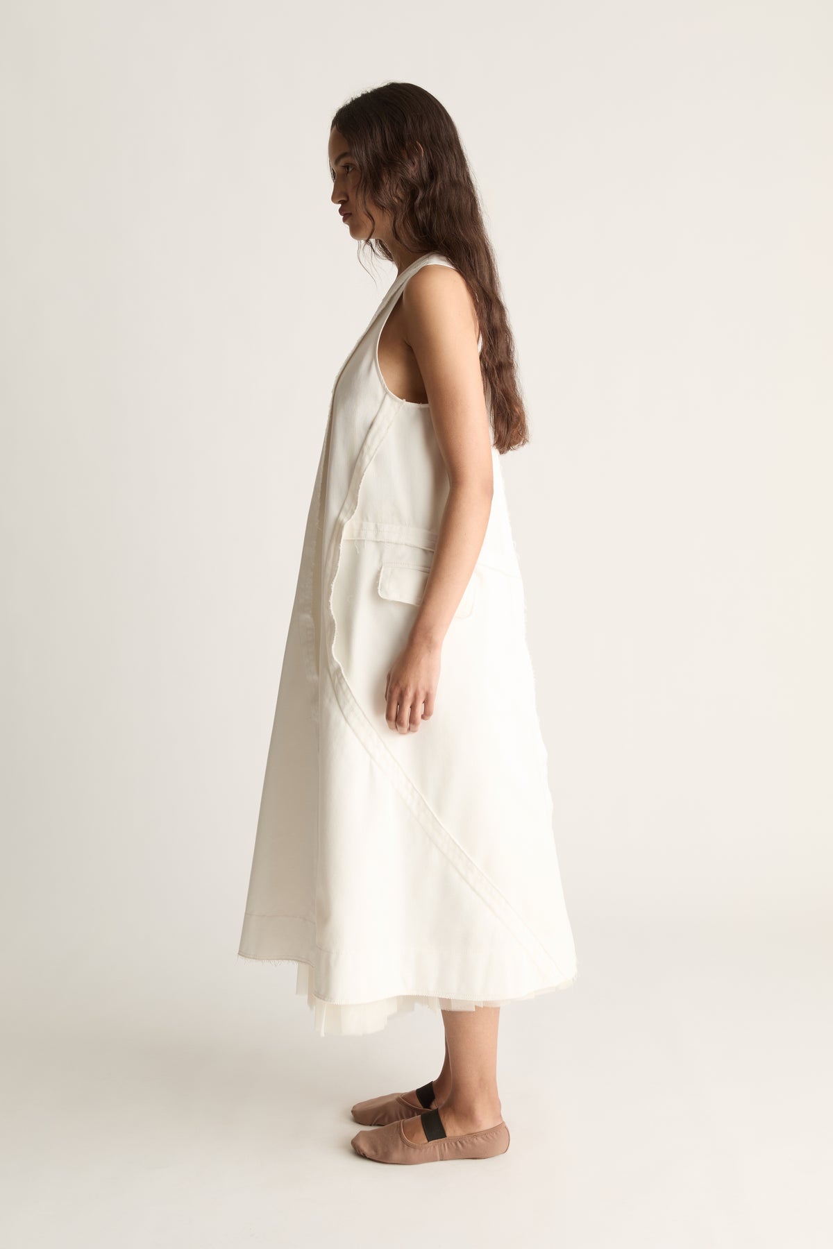 Side profile of a woman looking down in a white A-line dress with flap pocket, tulle hem and pink-beige flats
