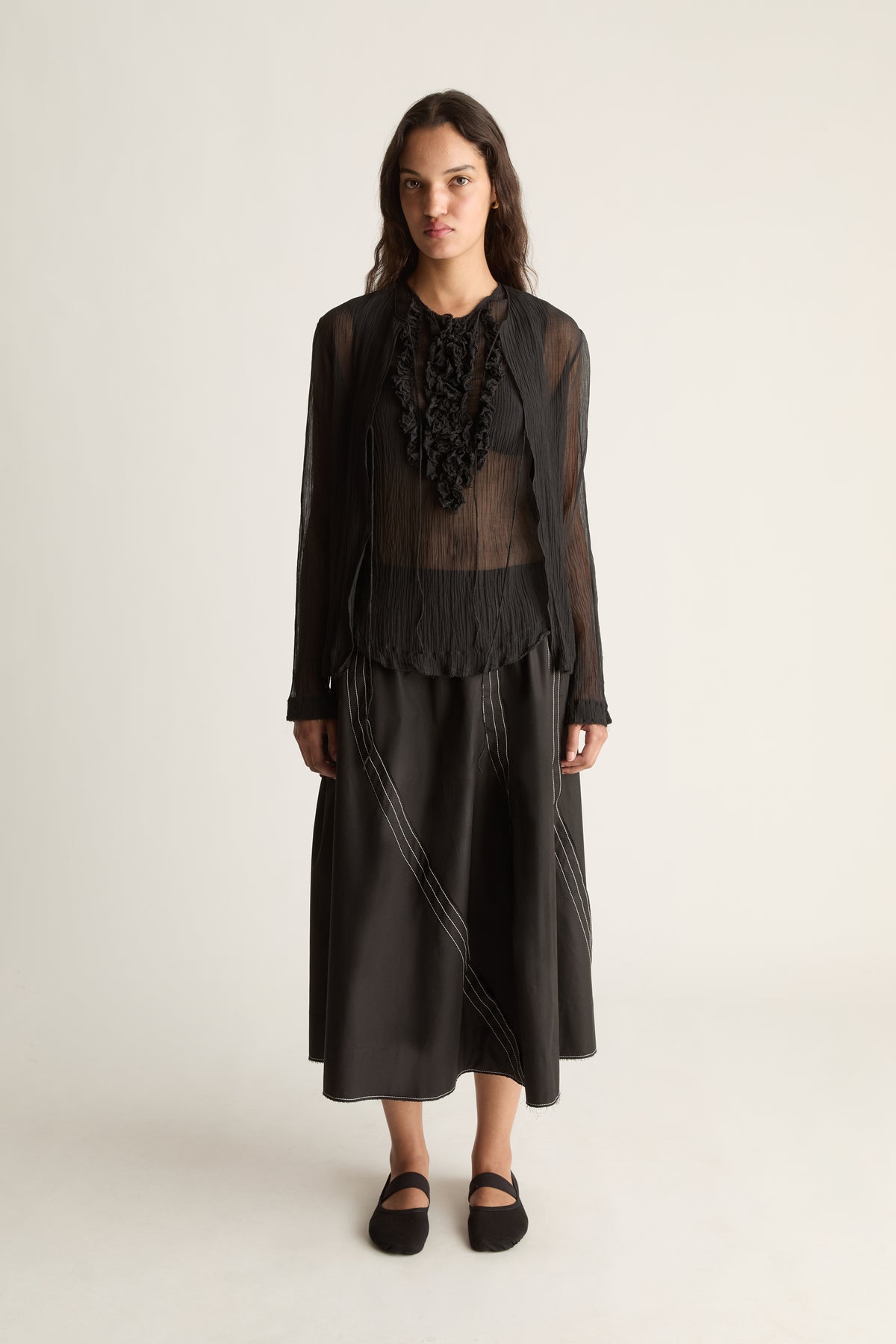 Front view of a woman wearing a sheer black cardigan, ruffle top and black skirt against a white studio background