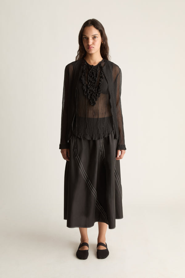 Front view of a woman wearing a sheer black cardigan, ruffle top and black skirt against a white studio background