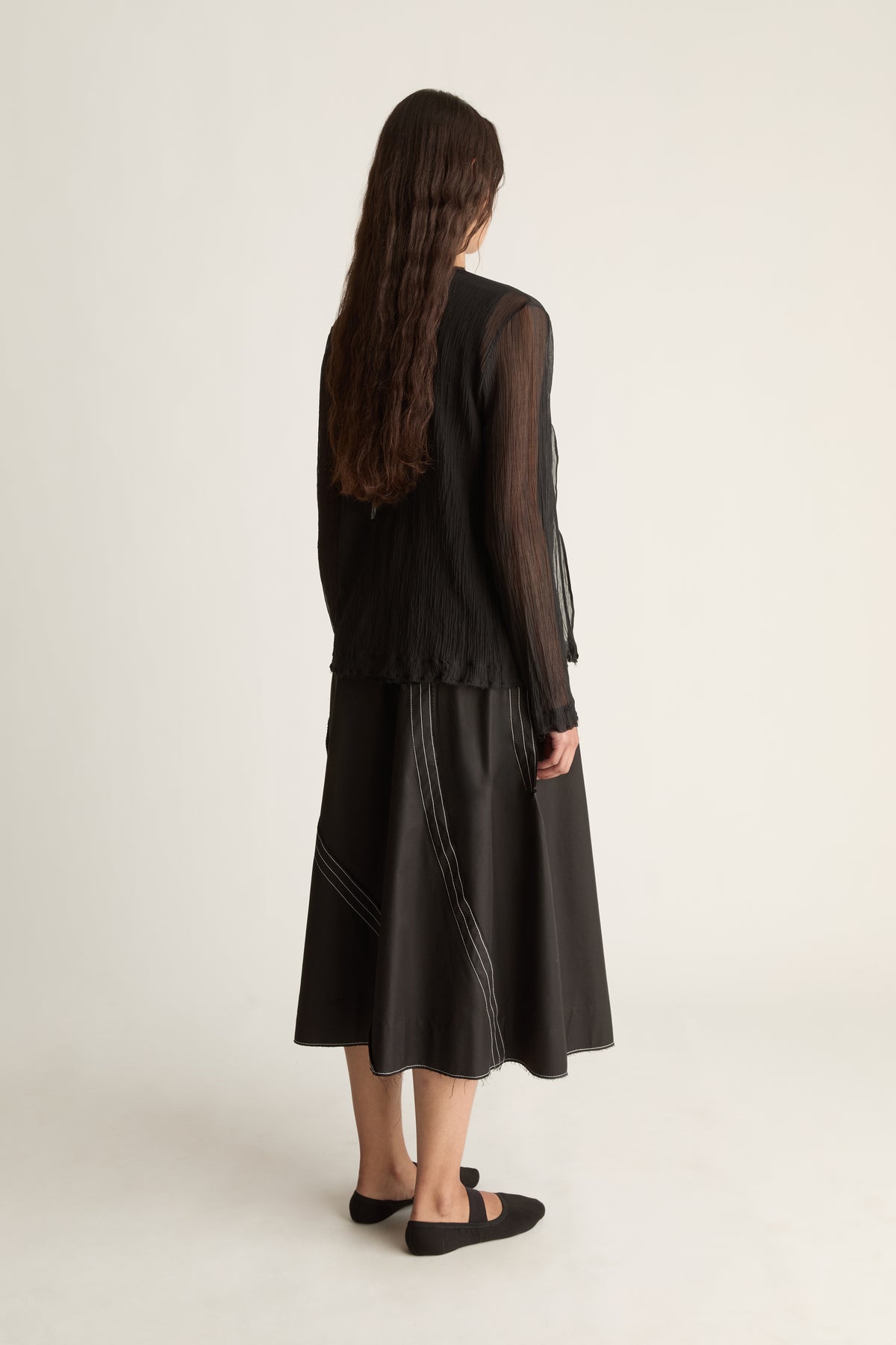 Back view of a woman with long wavy hair in a sheer black cardigan and black midi skirt