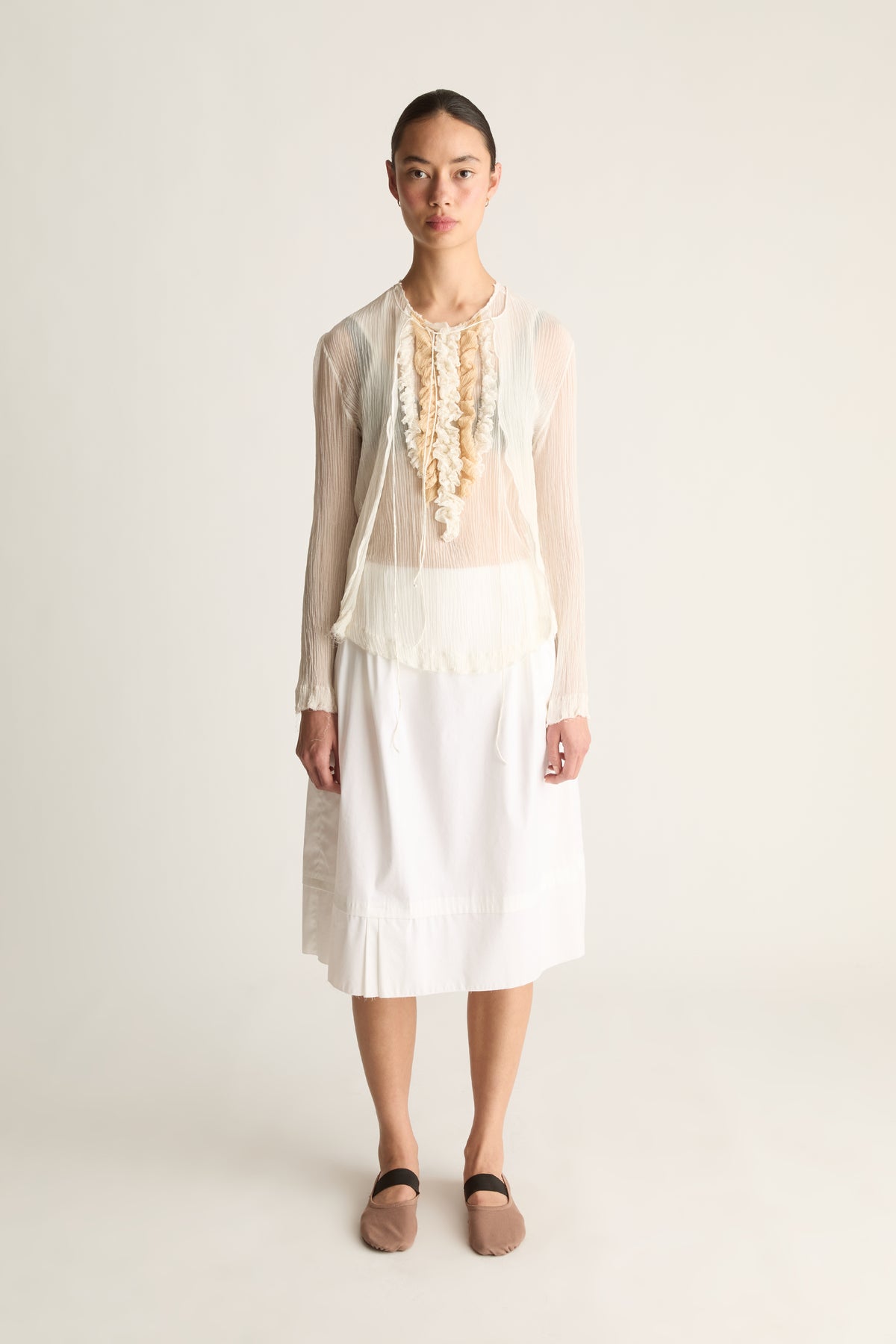 Front view of a woman wearing a sheer cream cardigan, ruffle top and white skirt against a white studio background