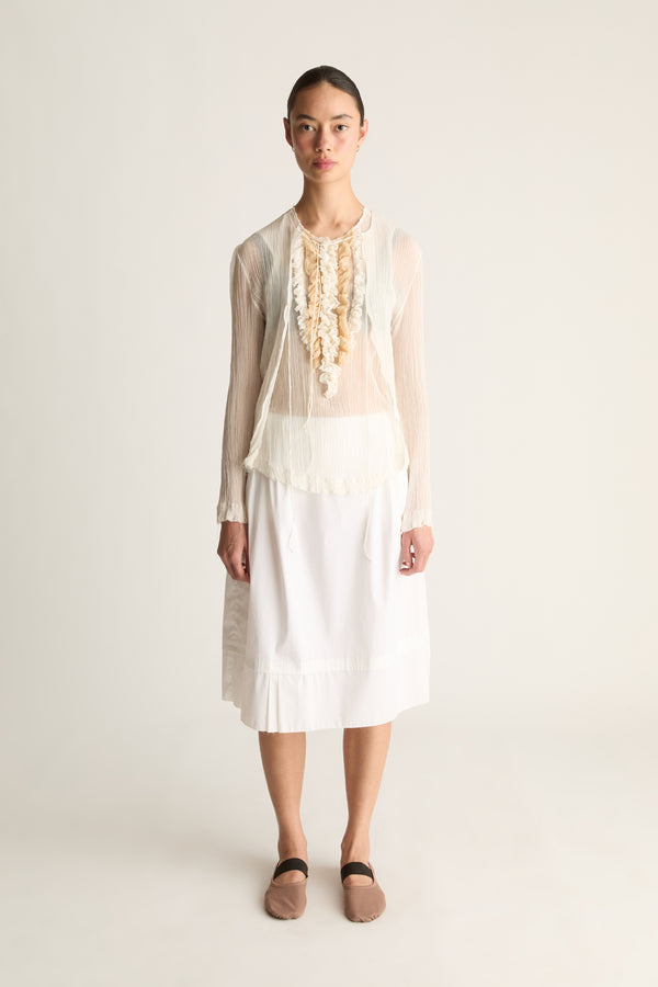 Front view of a woman wearing a sheer cream cardigan, ruffle top and white skirt against a white studio background