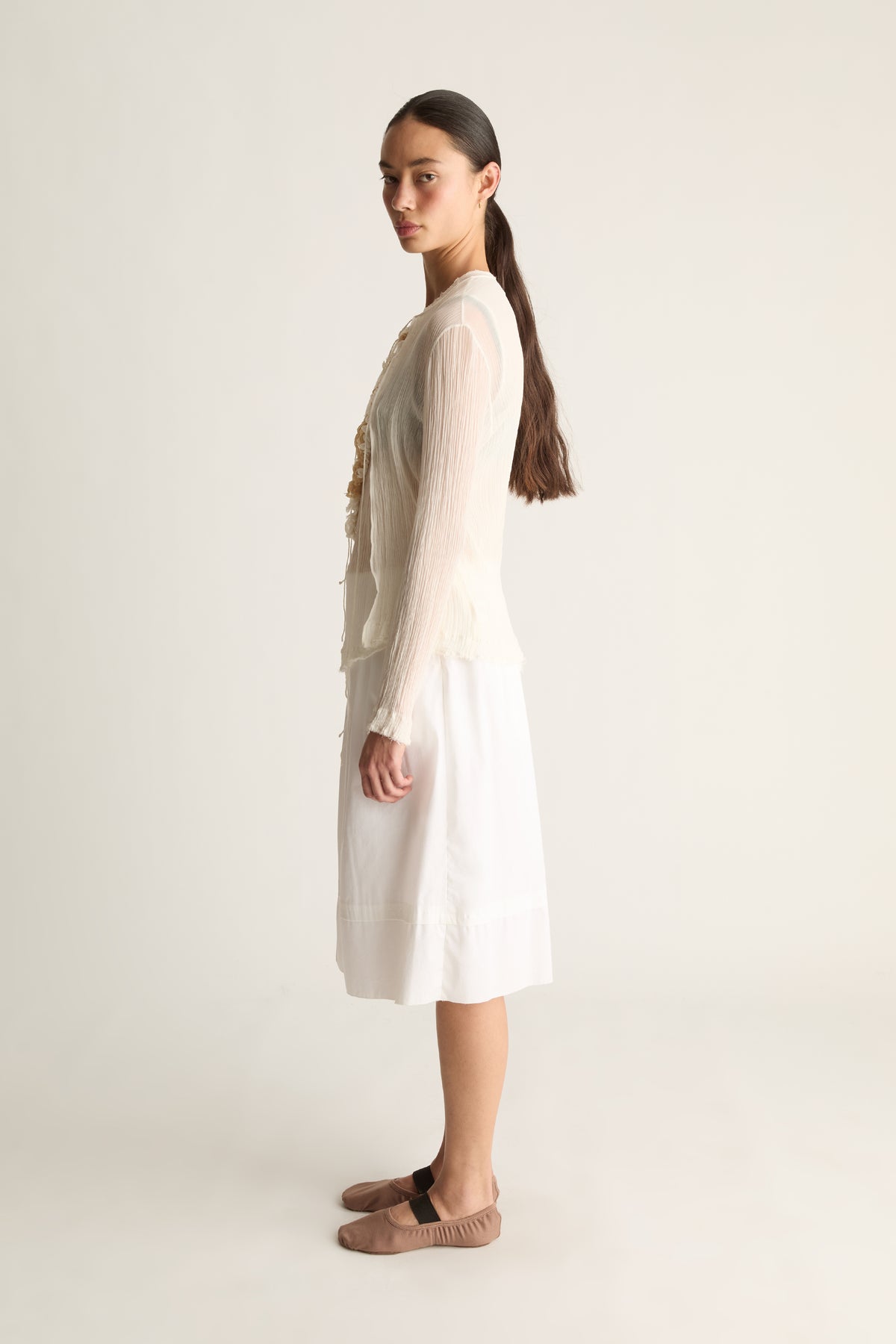 Side profile of a woman with a ponytail in a sheer cream long-sleeve cardigan and white skirt with taupe ballet flats