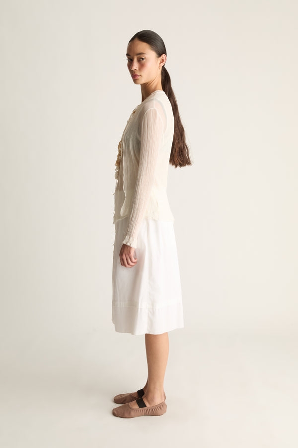 Side profile of a woman with a ponytail in a sheer cream long-sleeve cardigan and white skirt with taupe ballet flats