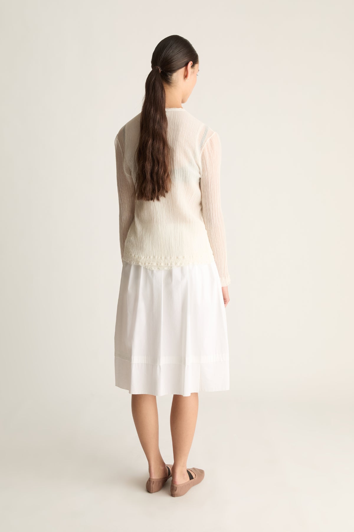 Back view of a woman with a ponytail in a sheer cream long-sleeve cardigan and a white midi skirt