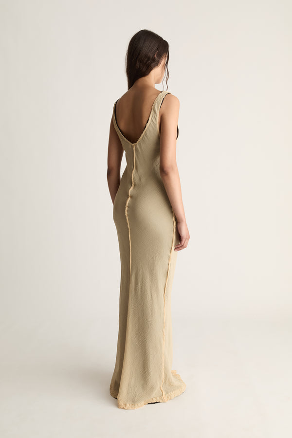 Back view of a woman with long dark hair in a beige maxi dress with low scoop back and raw-edge centre seam