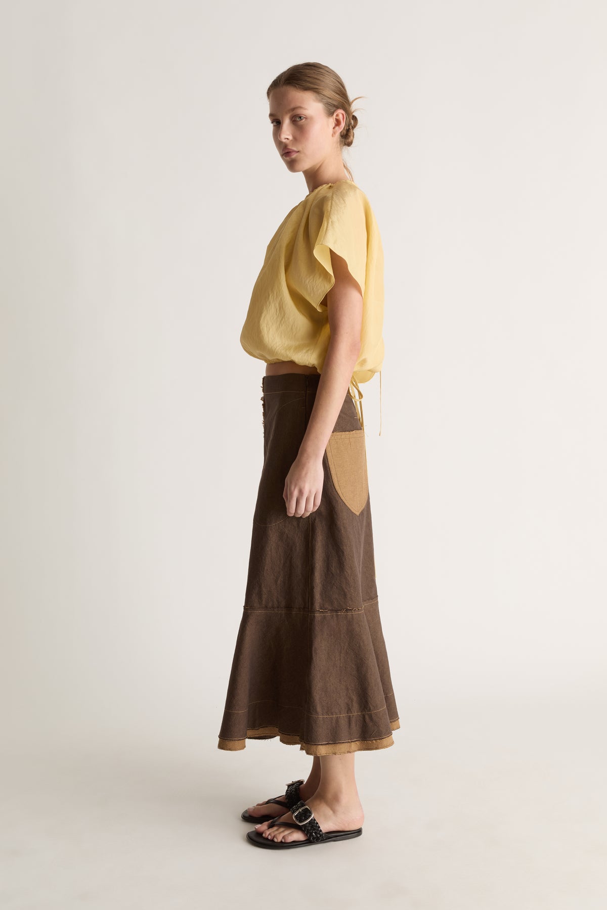 Side profile of a woman in a yellow short-sleeve gathered top and brown midi skirt with tan side pocket