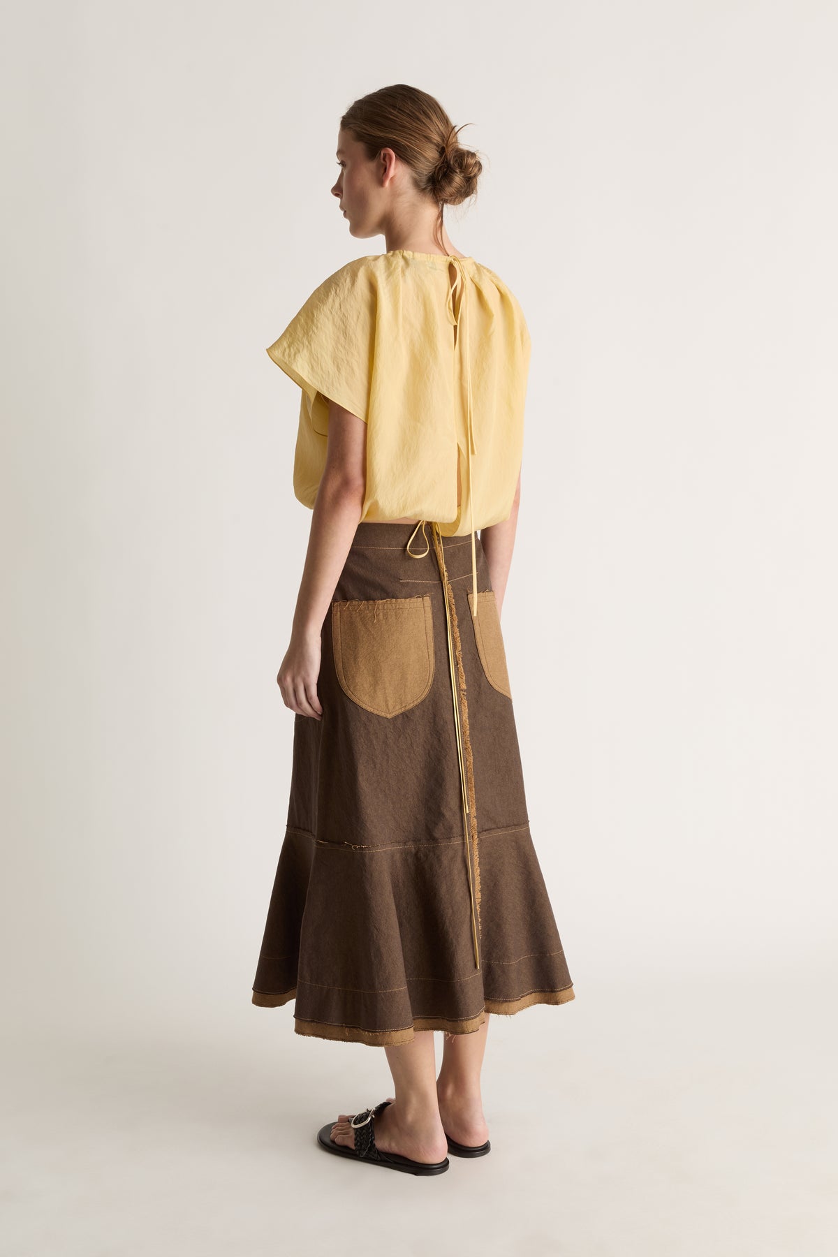 Back view of a woman with a bun in a yellow top with tie closure and a brown flared skirt with tan patch pockets