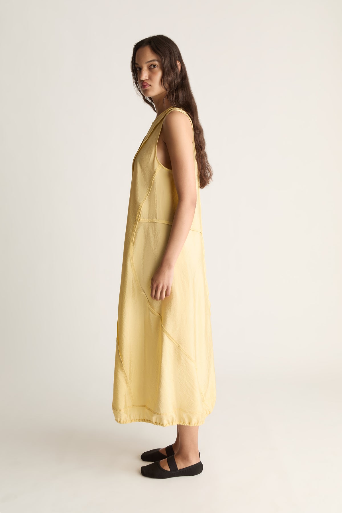 Side view of a woman looking over her shoulder in a yellow sleeveless midi dress and black ballet flats
