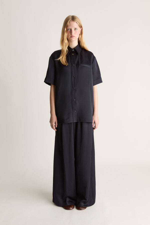 A woman stands against a plain light beige studio background wearing a dark navy short-sleeve button-front shirt with a chest pocket and matching dark wide-leg pants.