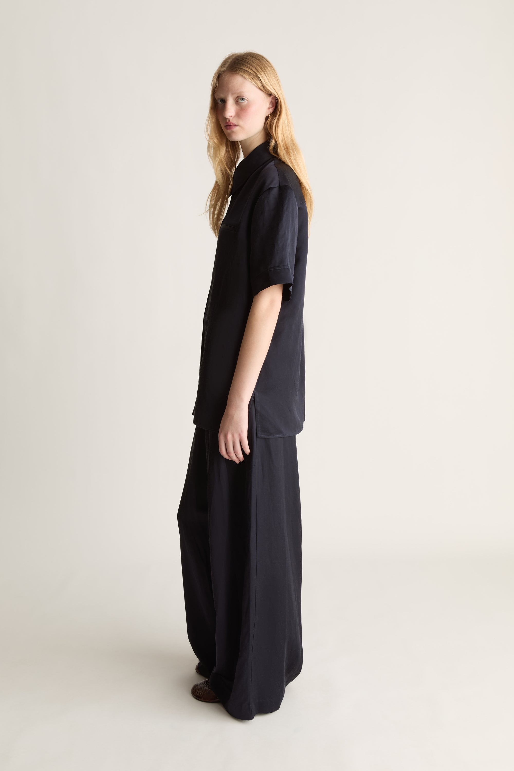 Side view of a woman modeling a navy short-sleeve collared shirt and matching wide-leg trousers against a plain light beige studio background.