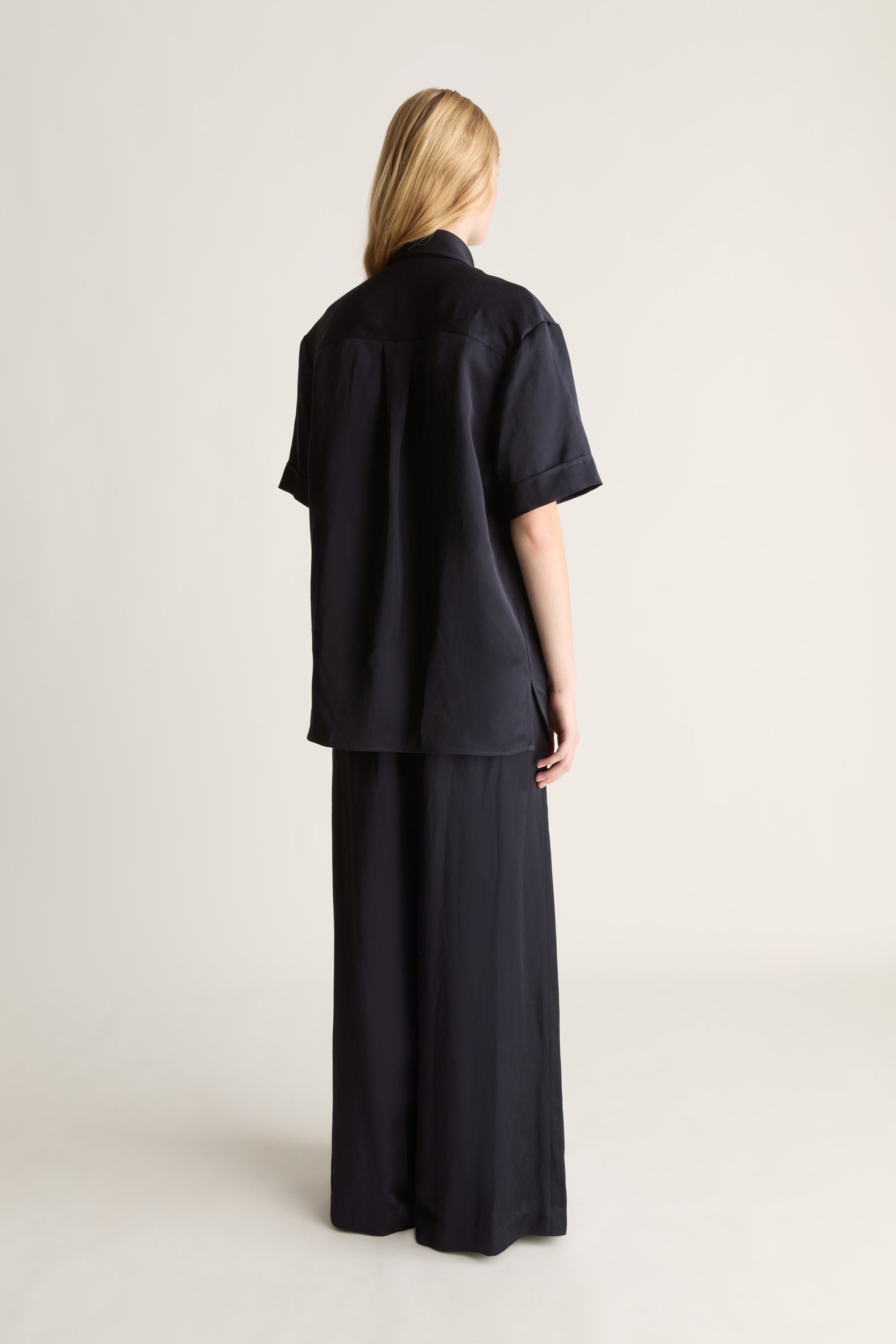 Back view of a woman wearing a navy short sleeve button up shirt and wide leg trousers, shot against a white studio background. 