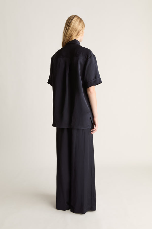 Back view of a woman wearing a navy short sleeve button up shirt and wide leg trousers, shot against a white studio background. 