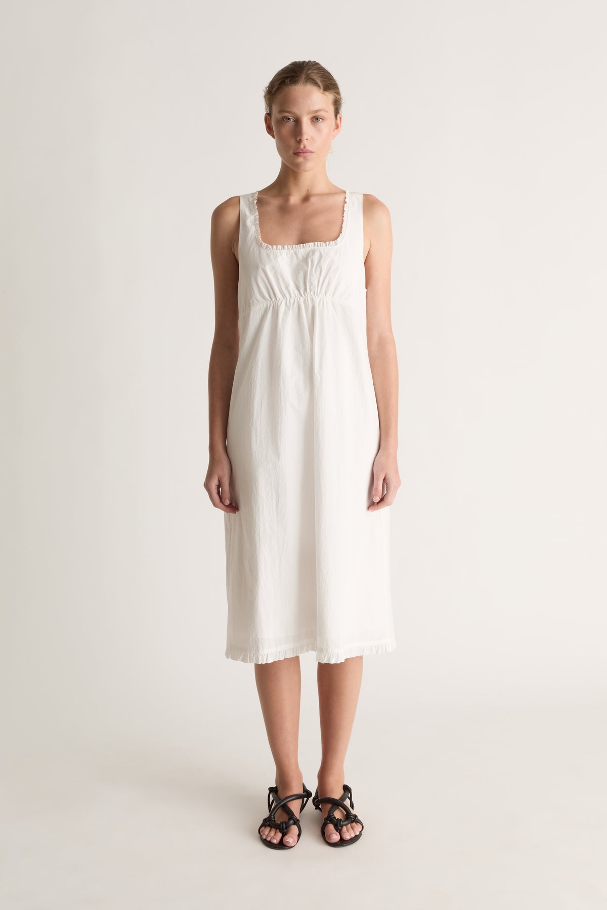 Front view of a woman wearing a white sleeveless midi dress with square neckline and ruffle trim against a white studio background