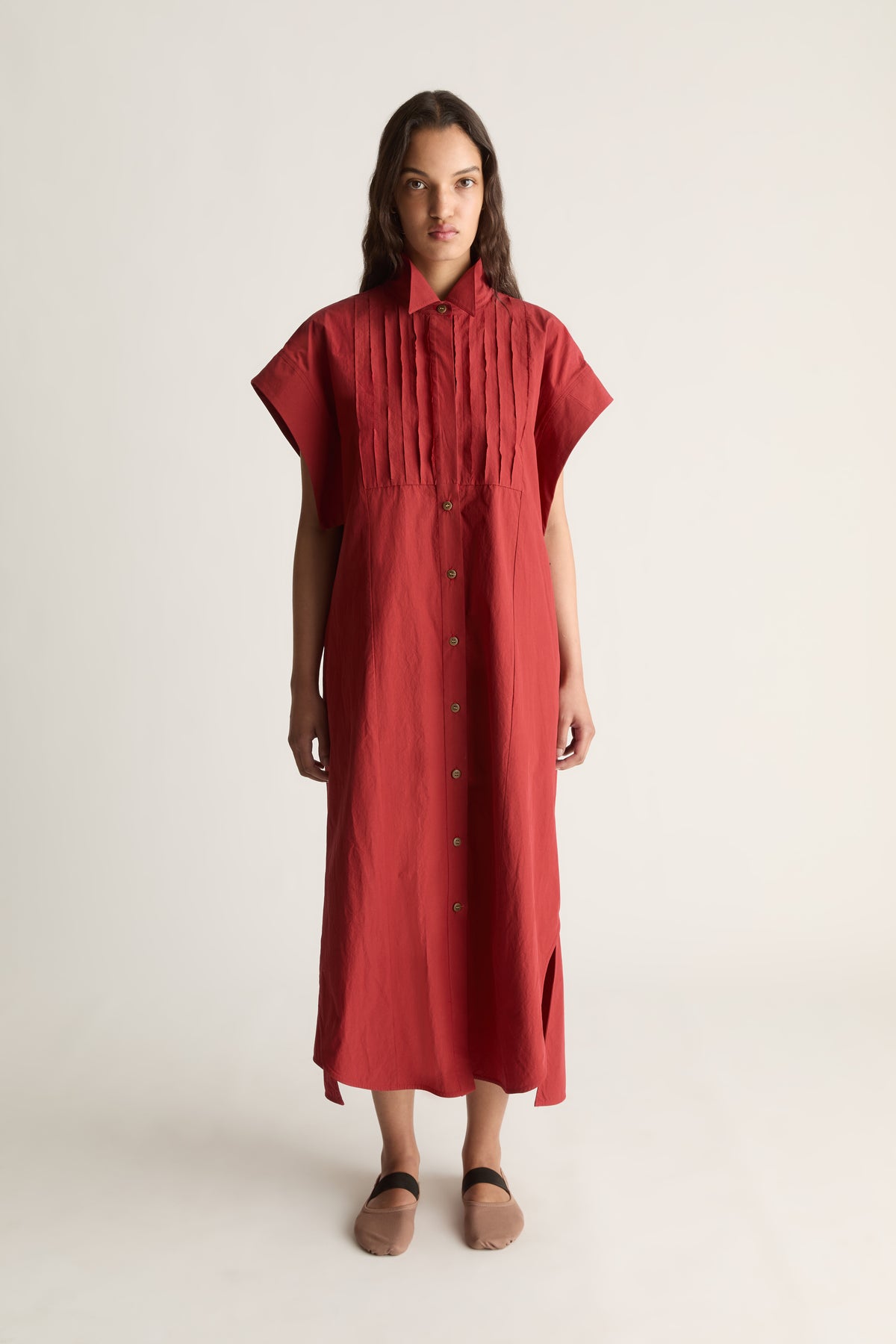 Front view of a woman wearing a red short-sleeve midi shirt dress with pleated bib and tan flats against a white studio background
