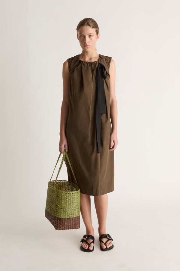 Front view of a woman in an olive-brown shift dress holding a green woven basket, with black sandals