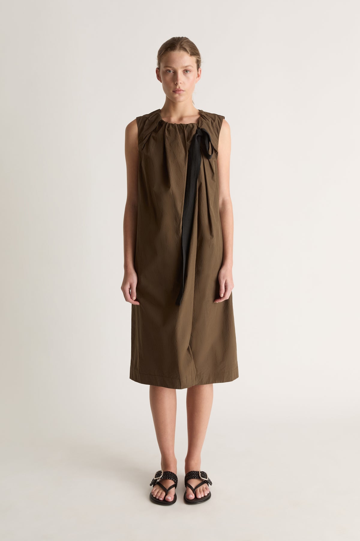 Front view of a woman wearing a khaki sleeveless midi dress with black ribbon tie against a white studio background