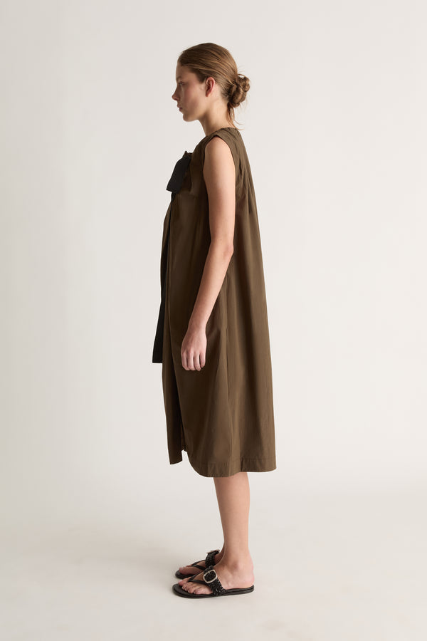 Side profile of a woman in a sleeveless khaki dress with black bow at the shoulder and black sandals